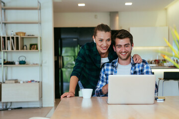 A young married couple is talking to parents, family and friends on a video call via a laptop while sitting in the living room of their modern house.