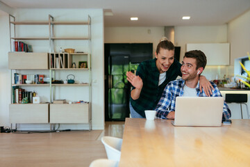 A young married couple is talking to parents, family and friends on a video call via a laptop while sitting in the living room of their modern house.