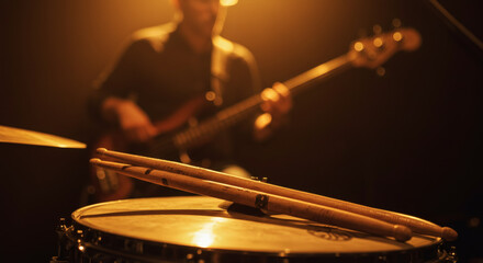 Drumsticks resting on snare drum with bassist in background atmospheric stage lighting live music performance concept