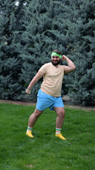 Vertical mobile street day photo. Warming up. Full body young adult white fat man with beard, fat belly in blue shorts, green headband, t-shirt, yellow sport sneakers raises leg to elbow in city park