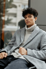 Fashionable young man in grey coat and turtle neck sweater sitting on bench, looking at phone in...