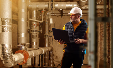 Engineer in a hard hat inspects industrial pipes in a boiler room, using a laptop to monitor...
