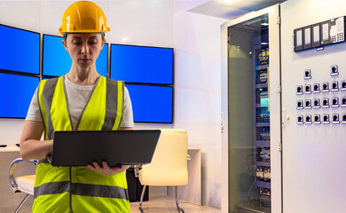 Engineer in a yellow vest and hard hat uses a laptop in a control room with servers and monitors. Scene relates to electrical engineering and system monitoring.