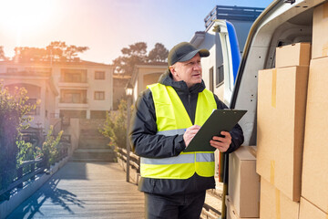 Delivery worker in a reflective vest checks a clipboard next to a van filled with packages in a...