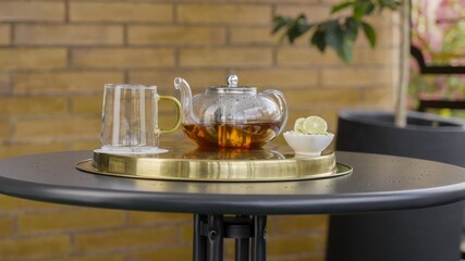 Transparent teapot and cup filled with black tea on a brass tray, with a bowl of lemon slices, set on a round outdoor table