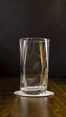 Clear empty glass with a unique twisted design placed on a white coaster atop a wooden table, dark background