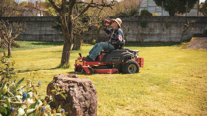 Man mowing spring lawn on red tractor in garden with apple and pear trees. Wearing work boots and flannel in rural setting.