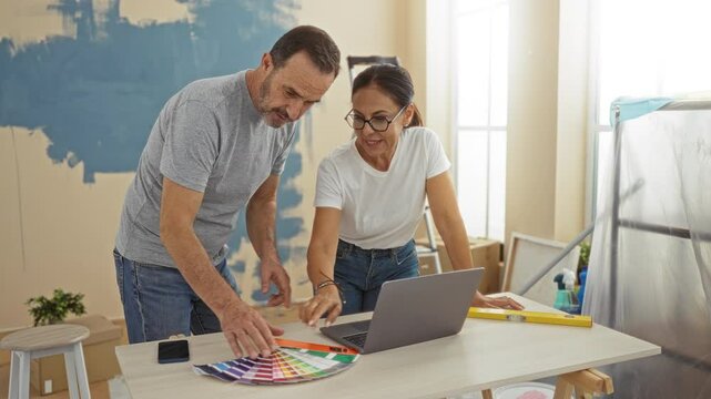 Middle-aged couple planning home renovation in new apartment, surrounded by paint samples and laptop, working together indoors with focus on interior design.