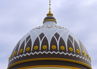 Islamic mosque dome with geometric arabesque pattern in gold, white, and brown against cloudy sky, showcasing traditional Muslim architecture and cultural heritage.

