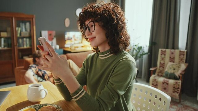 Young curly-haired woman checking social media on phone and smiling, sitting at table with coffee and snacks in sunny living room with boyfriend lounging on couch in background