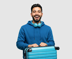 Smiling young male student in his mid-20s in blue hoodie, headphones around neck, holding travel suitcase, ready to fly for holiday, isolated on solid light background. 