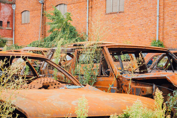 Carcasse de voitures abandonn&eacute;es. V&eacute;hicules br&ucirc;l&eacute;s. Casse automobile. Squelettes de voitures rouill&eacute;es devant une vieille usine de briques rouges. Cimeti&egrave;re de voitures