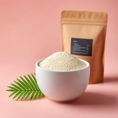 A bowl filled with a fine powder sits beside a brown packaging bag against a soft pink background, complemented by a green leaf for a fresh touch.