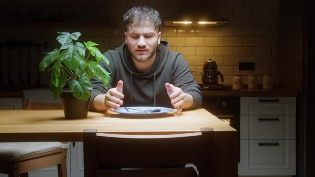 Hungry man sitting at kitchen table waiting for food with empty plate and cutlery in hand