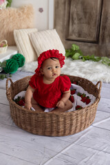 Cute baby girl in a red outfit and bonnet sitting in a wicker basket, holding and eating a strawberry, surrounded by fresh strawberries
