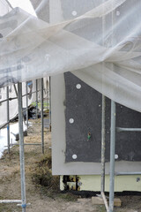 A wall of a house covered with EPS graphite polystyrene boards for thermal insulation, a scaffolding and building safety scaffolding net, some adhesive, anchors, an exterior wall mounted water faucet