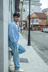 A young man wearing a shirt and jeans standing in front of a wall with his hands in his pockets, presenting a casual and confident appearance.
