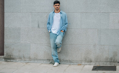 A young man wearing a shirt and jeans standing in front of a wall with his hands in his pockets,...