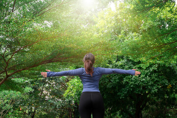 Rear view of a woman in sportswear stretching her arms wide in a green park, enjoying fresh air and...