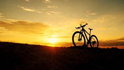 Obraz premium Silhouetted Mountain Bike at Sunset: A silhouette of a mountain bike stands on a hilltop, casting a bold silhouette against the fiery orange hues of a breathtaking sunset.