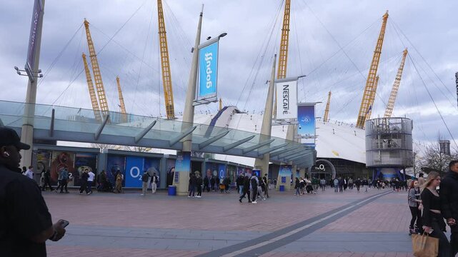 The O2 Arena in London is shown with its yellow masts and white dome. Pedestrians move around the plaza under an overcast sky, with banners visible.