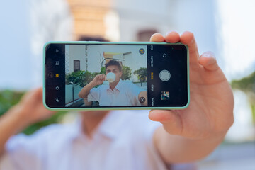 Young man holding smartphone at arms length taking a selfie outdoors. The phone screen shows his smiling face. A creative capture of mobile photography, connection, and personal expression.Vertical