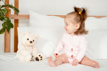 A baby girl in a pink bodysuit on the bed in the bedroom smiling or laughing, a cute little child at home with a teddy bear, a place for text