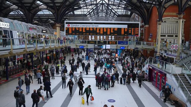 Dynamic scene of people walking and standing inside London Liverpool Street Station, with its arched roof, departure boards, and upper level shops.