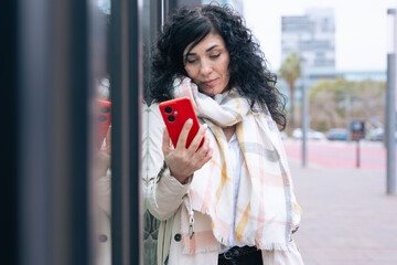 Fototapeta premium Happy caucasian woman standing outdoors in urban setting, wearing coat and scarf, using her red smartphone. She is smiling and relaxed, enjoying a casual moment on a cold day in the city. 