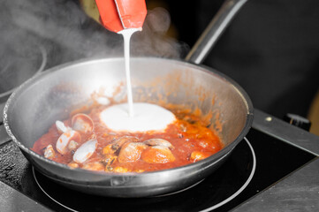 Creamy sauce is being poured into a pan of bubbling tomato sauce with clams. The steam rising...