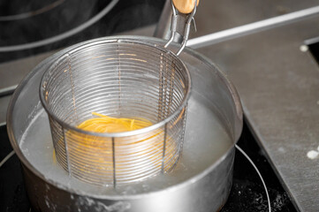 A wire mesh pasta strainer sits in a pot of boiling water, with strands of spaghetti visible...