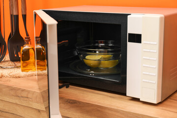Open microwave oven with bowl of water and lemons on wooden table indoors