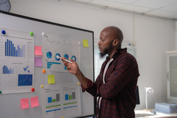 Businessman is pointing at a whiteboard with various financial charts and graphs, using a pen to highlight key data points during a presentation in a modern office setting