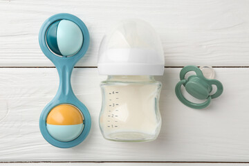 Feeding bottle with milk, pacifier and baby rattle on white wooden table, flat lay