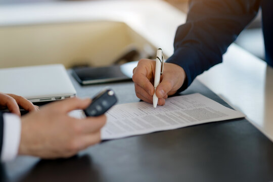Closeup shot of customer signing contract and salesman holding car keys in a dealership center, unrecognizable man buying vehicle in auto showroom, filling documents, cropped