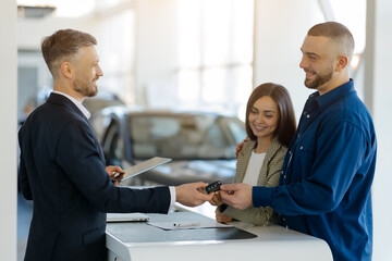 Happy caucasian millennial couple receiving car keys from salesman at a dealership showroom, young spouses buying new vehicle from auto salon, man and woman enjoying their purchase