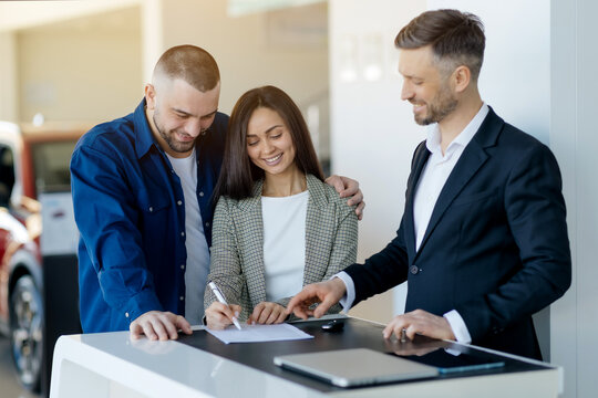 Happy young couple finalizing car purchase at dealership center, signing the contract. Salesman in black suit helping them through the process, showing where to sign, closeup