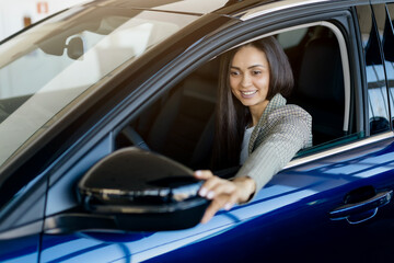 Millennial woman adjusting car side mirror while sitting inside new car in dealership showroom, happy young female enjoying driving, celebrating purchase new vehicle, closeup