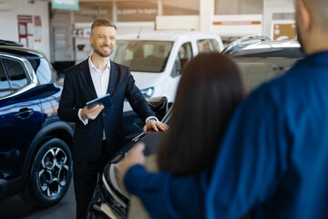 Smiling car salesman discussing vehicle options with a couple in the dealership showroom, man holding digital tablet and leaning casually against vehicle, advertising new auto