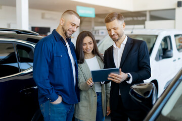 Salesman showing information about car on digital tablet to smiling couple, standing together...