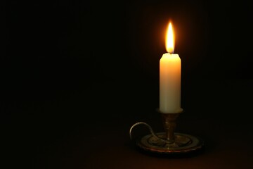 Burning candle in candlestick on black background, space for text