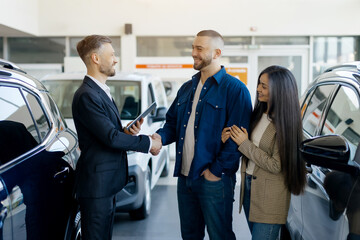 Friendly salesman shaking hands with a smiling couple inside a bright, modern car dealership...