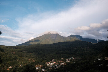Serene view of a lush green mountain partially covered by misty clouds with a small village nestled at its forested base, captured under soft evening light and a clear sky.