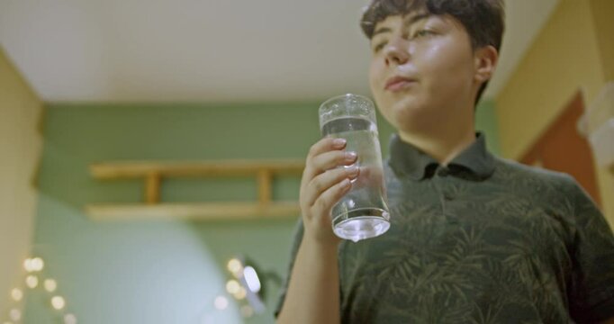 A girl helps her pet green-cheeked conure parrot to drink water from a glass in her home. The bird then perches on top of its cage by the window.