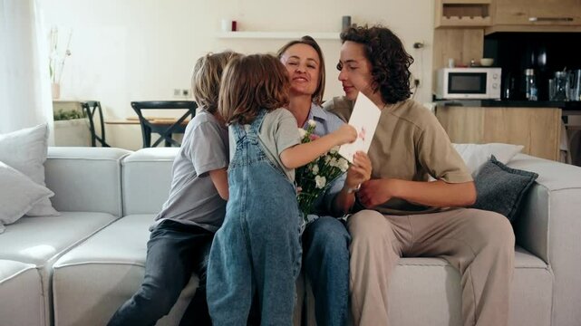 Three children celebrate Mother's Day by giving their mom flowers and a greeting card at home. She smiles, surrounded by love and joy. Concept of motherhood and family connection. Mid-range shot