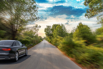 A black car speeds down a tree-lined country road at sunset, creating a dynamic motion blur through...