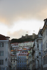 Obraz premium The medieval Castelo de São Jorge looms over a colourful street in Bairro Alto, Lisbon, its historic ramparts contrasting with the vibrant buildings below, capturing city’s rich heritage at sunrise.