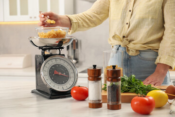 Woman adding raw pasta into bowl on mechanical kitchen scale at white marble table indoors, closeup