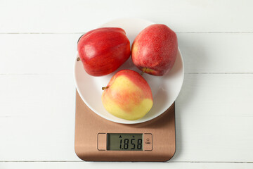 Modern electronic kitchen scale with apples on white wooden table, top view