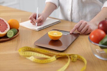 Woman weighting orange and writing in notebook at wooden table, closeup